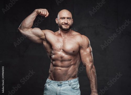 Portrait of a happy and muscular guy with bald head and naked torso posing in dark background showing his huge biceps looking at camera.