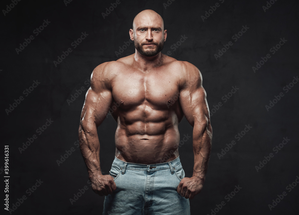 Muscular and bald caucasian man with perfect abs and huge biceps poses in dark background looking at camera with serious face.