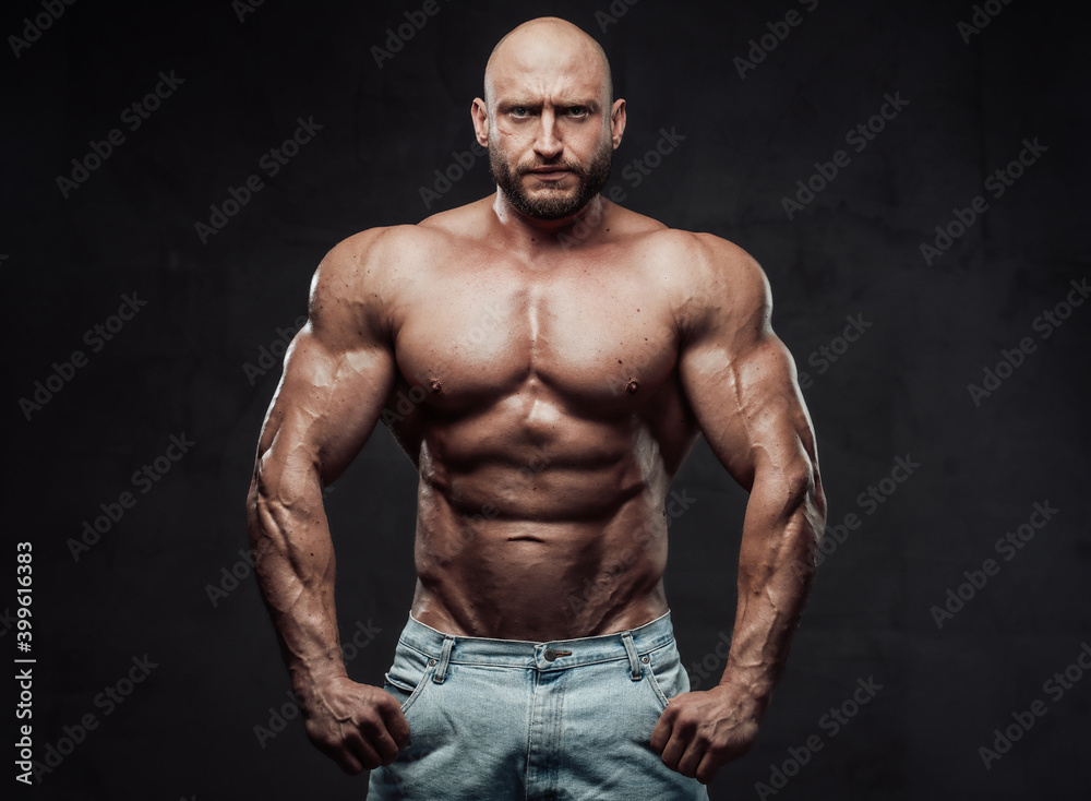 Bearded and hairless man with powerful and muscular build in jeans shorts poses with serious face in dark background looking at camera.