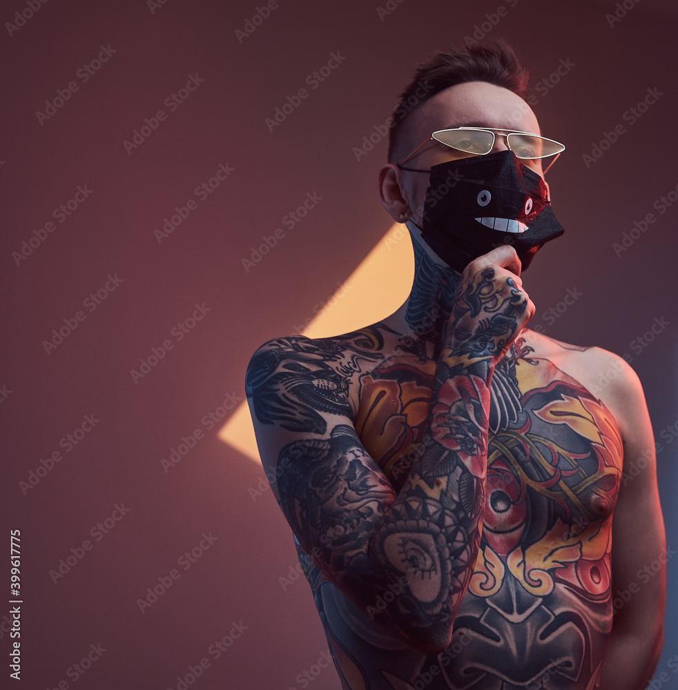Individual and tattooed macho weared with sunglasses and protective mask poses in dark studio room with spotlight and colourful walls. Individual and tattooed macho weared with sunglasses and protective mask poses in dark studio room with spotlight and colourful walls.