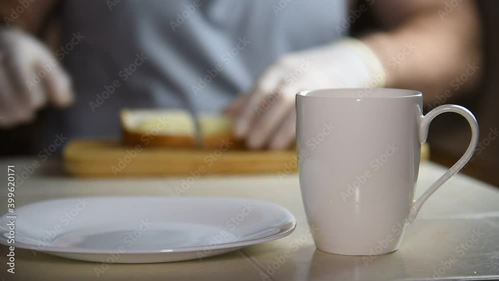A man cuts bread and butter, puts it on a plate, pours hot water into a mug, the background is blurred. Breakfast in the morning. A man cuts bread and butter, puts it on a plate, pours hot water into a mug, the background is blurred. Breakfast in the morning.