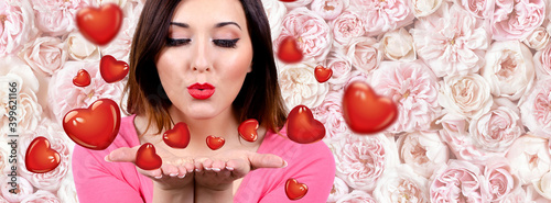 Valentine's day. A pretty woman blowing a kiss kisses in the shape of a heart. The background is a wall of pink flowers.  copy space.