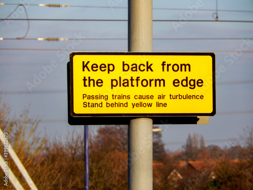 Yellow sign warning people to keep back from the edge of a railway station platform when trains are passing.