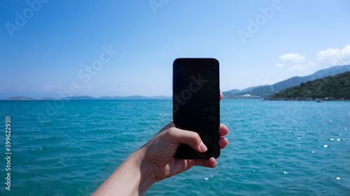 Fototapeta Naklejka Na Ścianę i Meble -  Vertical smartphone. Tourist hand holding a black blank space screen smart phone while in vacation in the beach with view of the water blue landscape. Coast of Turkey, Aegean Sea