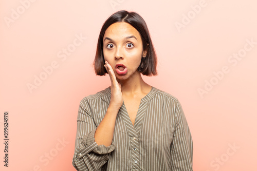 young hispanic woman feeling shocked and astonished holding face to hand in disbelief with mouth wide open photo