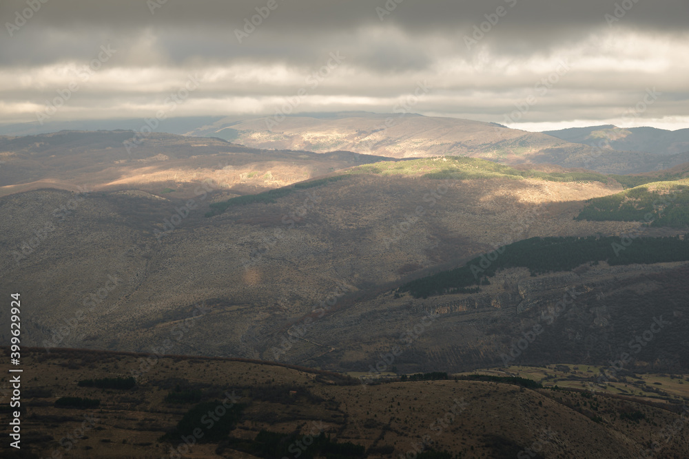 Beautiful patches of light on a hills and valleys of a rocky mountain fields covered by pine trees and dark, moody sky