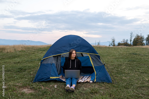 Girl with laptop in the mountains. Tourist woman uses a laptop in nature, freelancer typing on a computer near a tent in a journey against the backdrop of a mountain landscape.