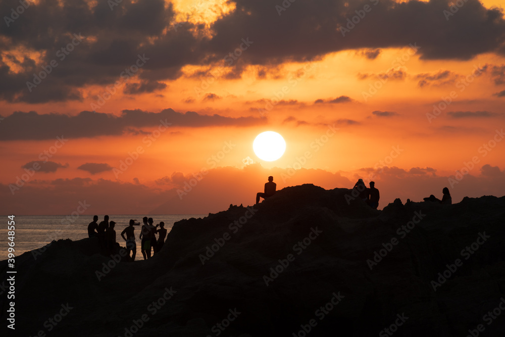 Sunset silhouettes. Groups of young people looking at the sunset from a rock in the sea on the coast of Tropea, Calabria. italian summer and vacation mood, amazing colored sunset sky, reflections.