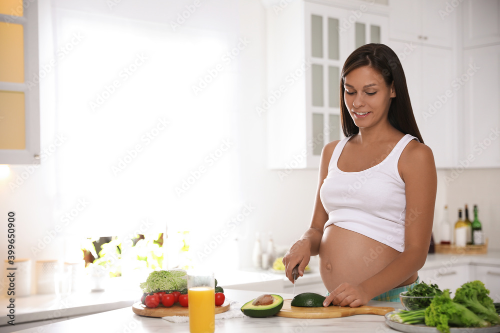 Young pregnant woman cutting avocado at table in kitchen. Taking care of baby health Young pregnant woman cutting avocado at table in kitchen. Taking care of baby health