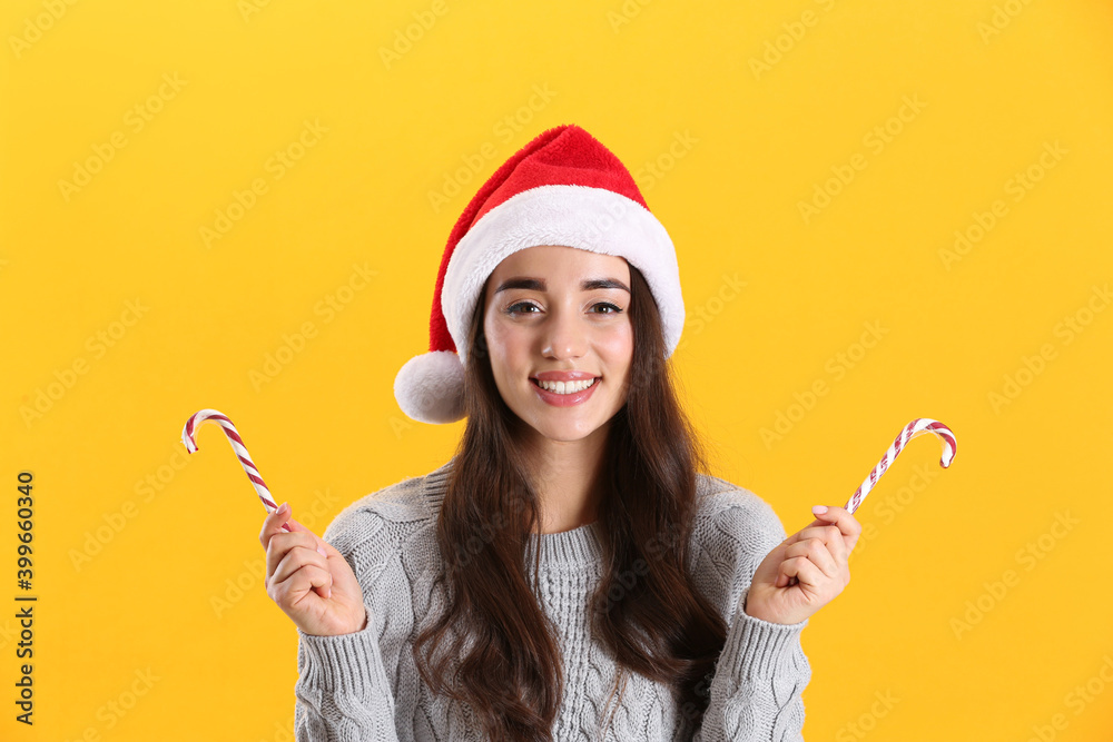 Beautiful woman in Santa Claus hat holding candy canes on yellow background Beautiful woman in Santa Claus hat holding candy canes on yellow background