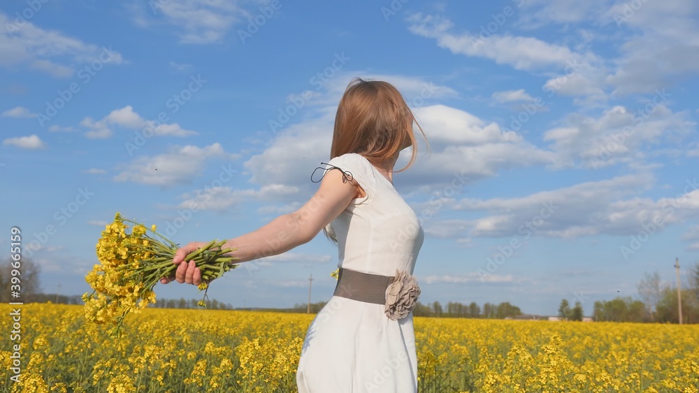Fototapeta premium Happy girl in a white dress with a bouquet of rapeseed flowers in a yellow rapeseed field.