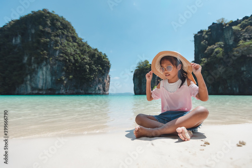 Cute asian child girl wearing hat and sunglasses sitting on the beach and playing with sand. She is enjoying with beautiful nature in her vacation with family.