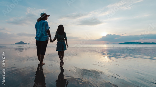 Silhouette of asian mother and daughter holding hand and walking on the beach together at the sunset time with beautiful sea and sky. Family enjoy with nature concept.