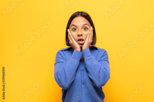 young latin woman looking unpleasantly shocked, scared or worried, mouth wide open and covering both ears with hands photo