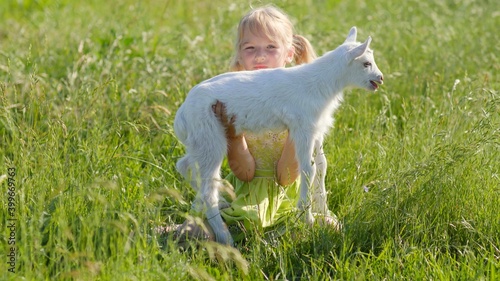 A six-year-old girl in a meadow gently hugs a small white goat.