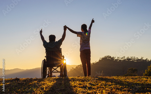 Disabled handicapped young man in wheelchair raised hands with his care helper in sunset.Silhouette