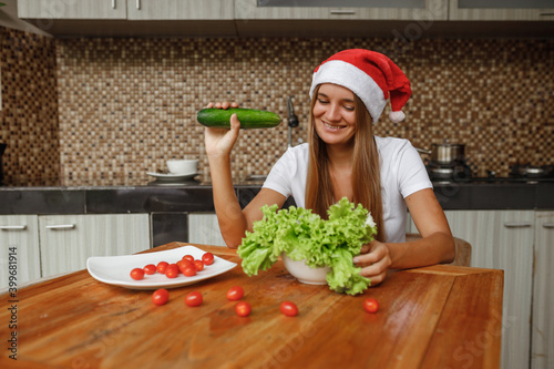 Smiling young woman in christmas hat sit on the kitchen at home and eat vegetable alad photo