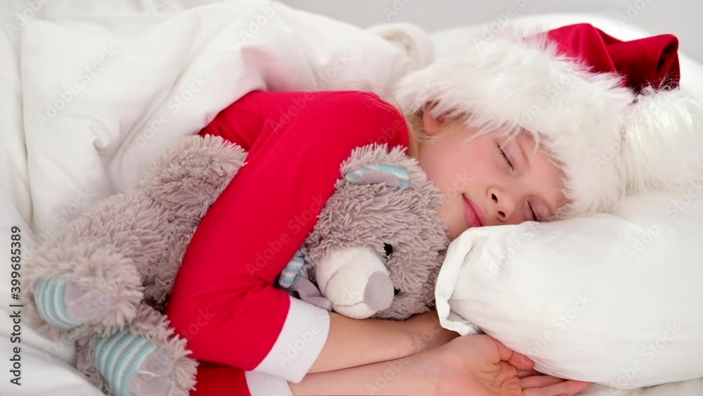 Child sleeping before Christmas. Little girl in Santa hat and Christmas pajamas lying in white bed hugging teddy bear at home. New Year and Holidays. morning happiness Child sleeping before Christmas. Little girl in Santa hat and Christmas pajamas lying in white bed hugging teddy bear at home. New Year and Holidays. morning happiness