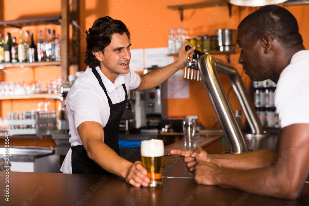Smiling man barman giving glass of golden beer to client in cafe
