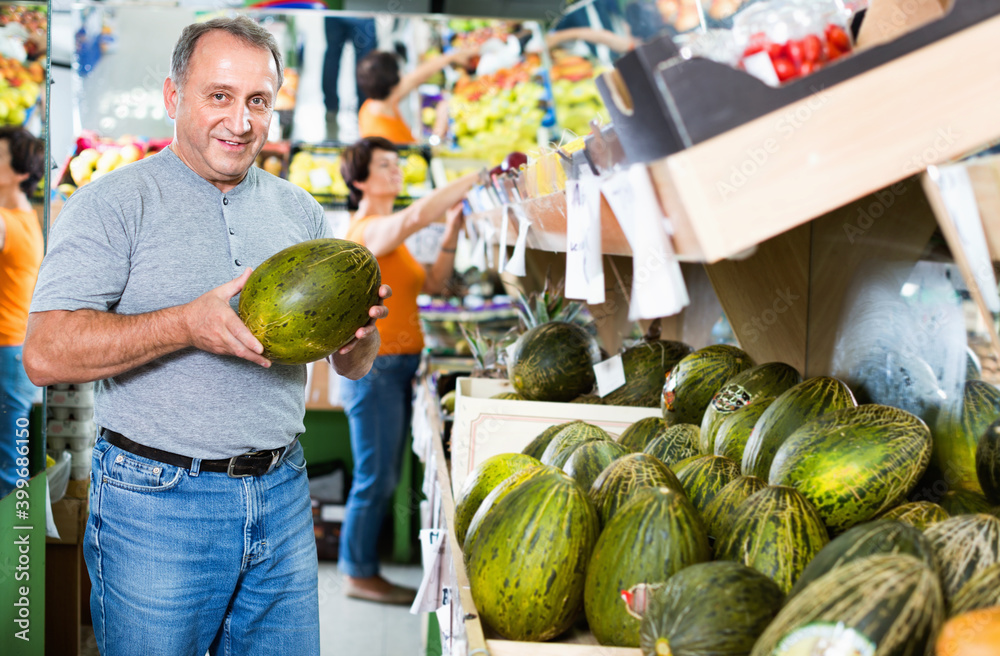 Fototapeta premium Smiling positive glad male is choosing green melon in the supermarket.