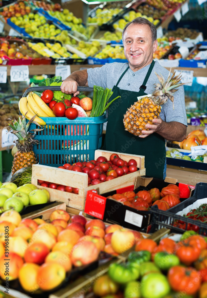 Smiling cheerful man seller is standing with basket with fruits and vegetables in the supermarket.