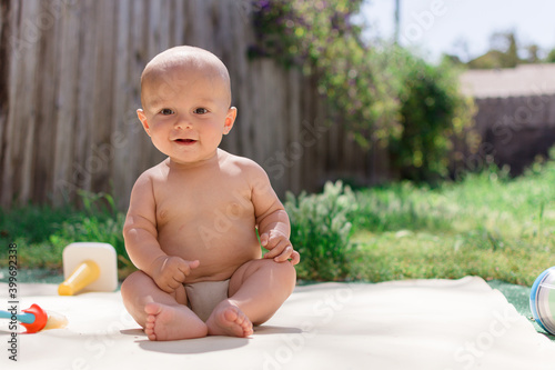 Bald baby with brown eyes sitting in a back yard with toys and a blurry background of grass and a fence photo