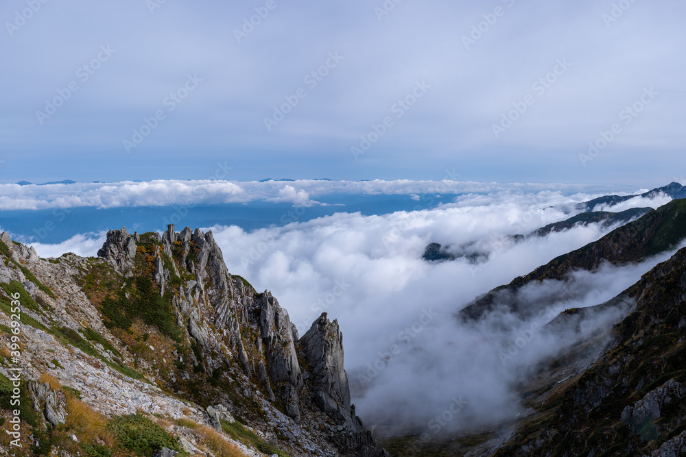 View of rocky mountain range and valley engulfed in clouds on the horizon in early autumn at Senjojiki Cirque in Nagano Prefecture, Japan. View of rocky mountain range and valley engulfed in clouds on the horizon in early autumn at Senjojiki Cirque in Nagano Prefecture, Japan.