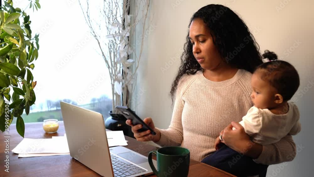 African American mother holds her mixed race infant daughter as she texts a message on her cell phone with one hand with laptop, mug and papers sitting on a temporary work from home dining table desk.