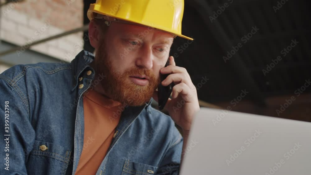 Close up shot of bearded male engineer in hardhat talking on mobile phone and typing on laptop while working at construction site Close up shot of bearded male engineer in hardhat talking on mobile phone and typing on laptop while working at construction site