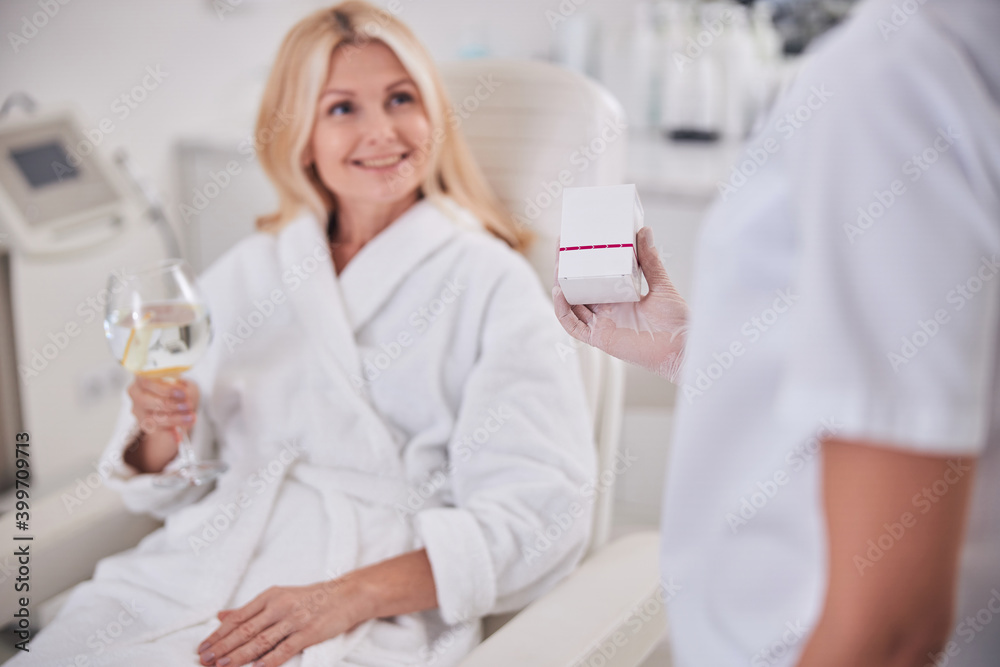 Blonde Caucasian female with glass if water in hand listening her doctor in cabinet Blonde Caucasian female with glass if water in hand listening her doctor in cabinet