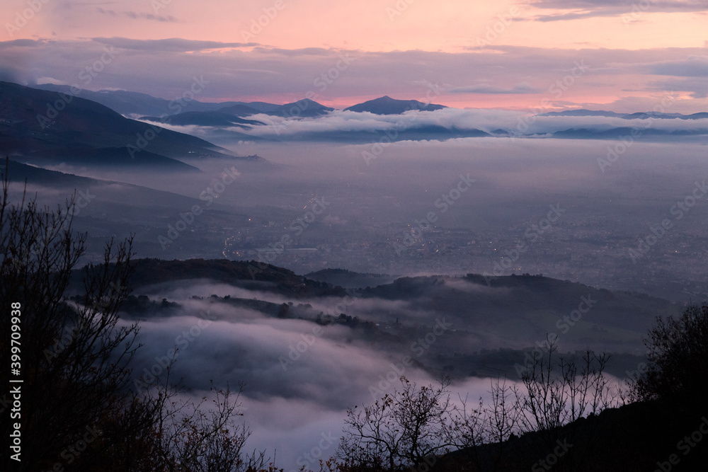Mist and fog between valley and layers of mountains and hills at dusk, in Umbria (Italy), with city lights underneath