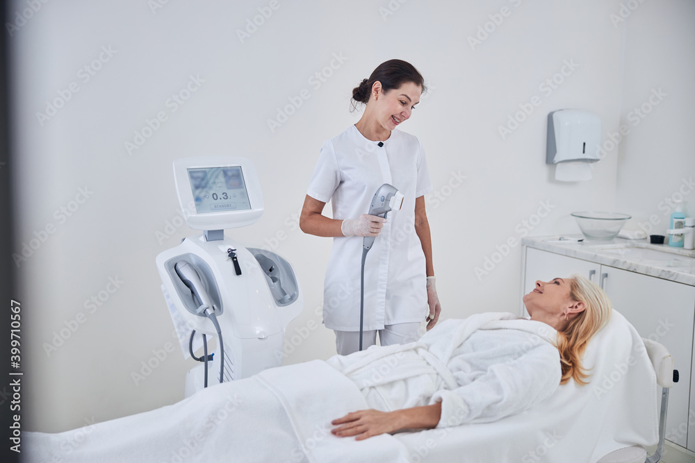 Brunette female cosmetologist holding ultrasound modern machine in hand in beauty salon Brunette female cosmetologist holding ultrasound modern machine in hand in beauty salon