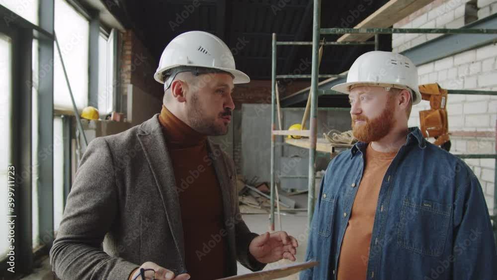 Arc waist up shot of architect and male client in helmets standing in new unfinished apartment and having discussion Arc waist up shot of architect and male client in helmets standing in new unfinished apartment and having discussion