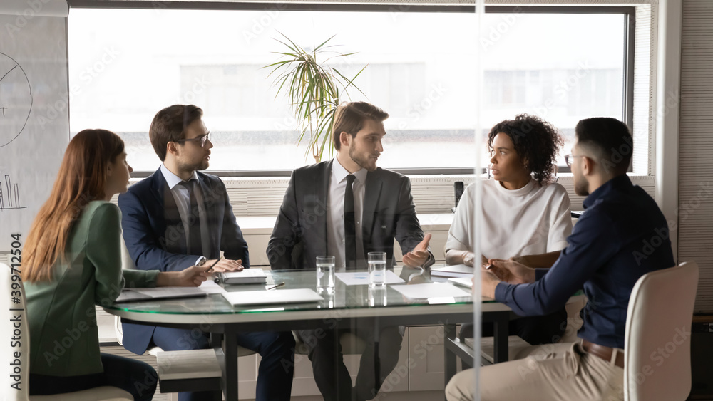 Young multiracial employees sit at table in office boardroom talk brainstorm discussing company business ideas together. Diverse multiethnic businesspeople engaged in team discussion at meeting.