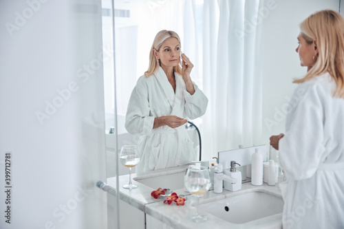 Seriously business woman in white plush bathrobe looking to the mirror in in bathroom in hotel