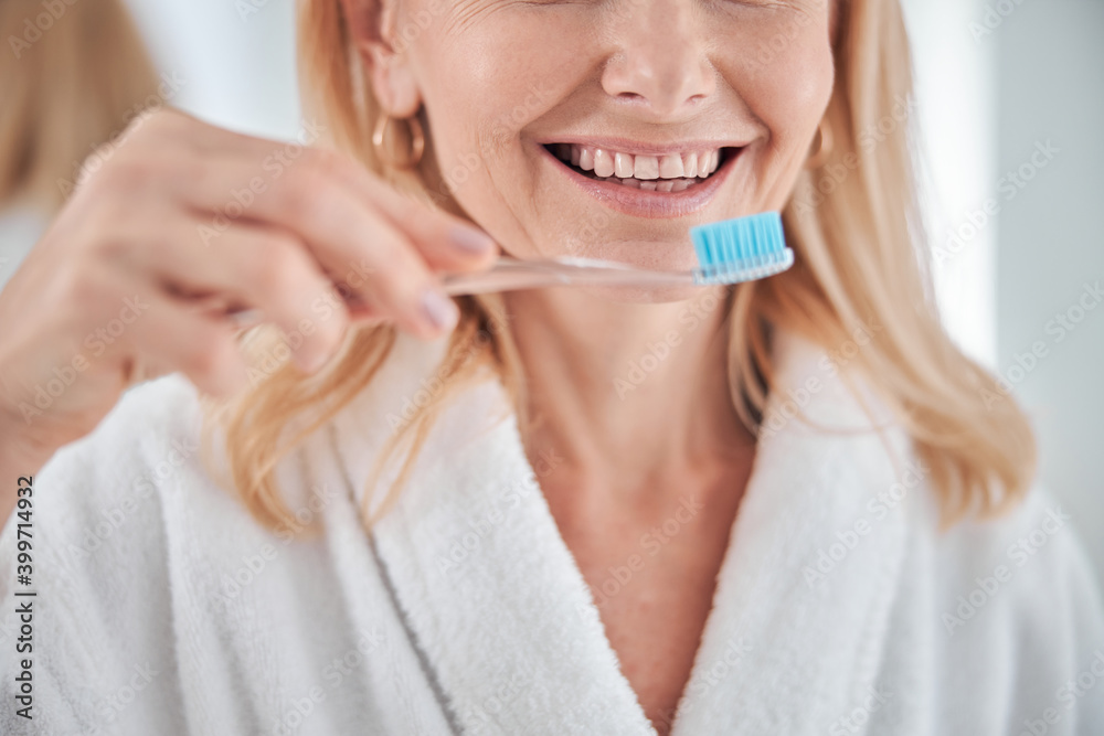 Beautiful cheerful blonde female in white plush bathrobe taking care for her teeth Beautiful cheerful blonde female in white plush bathrobe taking care for her teeth