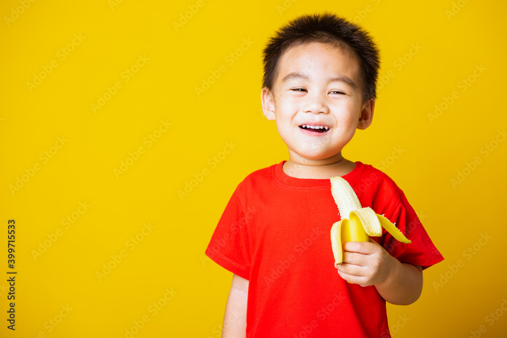Happy portrait Asian child or kid cute little boy attractive smile wearing red t-shirt playing holds peeled banana for eating, studio shot isolated on yellow background Happy portrait Asian child or kid cute little boy attractive smile wearing red t-shirt playing holds peeled banana for eating, studio shot isolated on yellow background