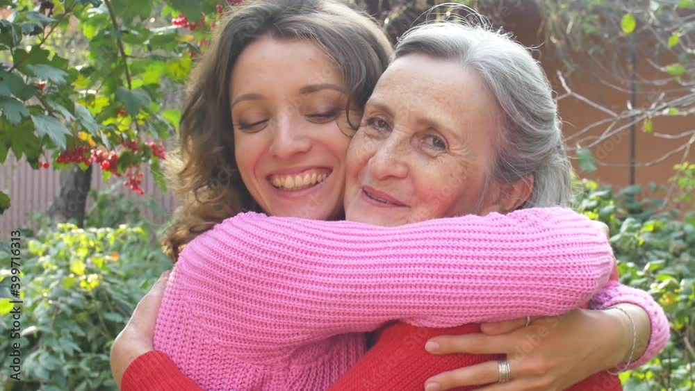 Senior mother with gray hair with her adult daughter looking at the camera in the garden and hugging each other during sunny day outdoors Senior mother with gray hair with her adult daughter looking at the camera in the garden and hugging each other during sunny day outdoors