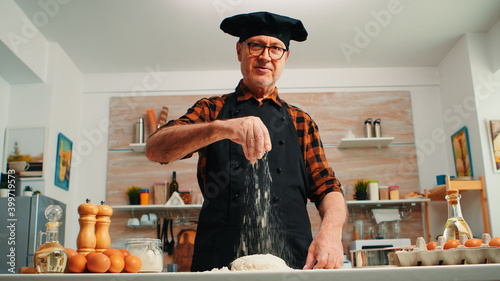 Senior man adding flour on dough by hand looking at camera smiling. Retired elderly chef with bonete and uniform sprinkling, sieving spreading rew ingredients with hand baking homemade pizza and bread photo