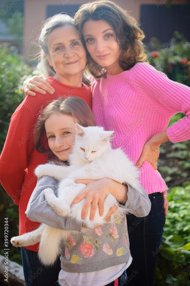 Senior mother with gray hair with her adult daughter and little granddaughter with white cat are looking at the camera in the garden and hugging each other during sunny day outdoors Senior mother with gray hair with her adult daughter and little granddaughter with white cat are looking at the camera in the garden and hugging each other during sunny day outdoors