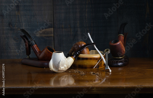 Composition with smoking pipes: in the foreground a pipe made of sea foam (meerschaum), two of heather and one of cherry, an ashtray, pipe cleaning accessories and tobacco crumbs.