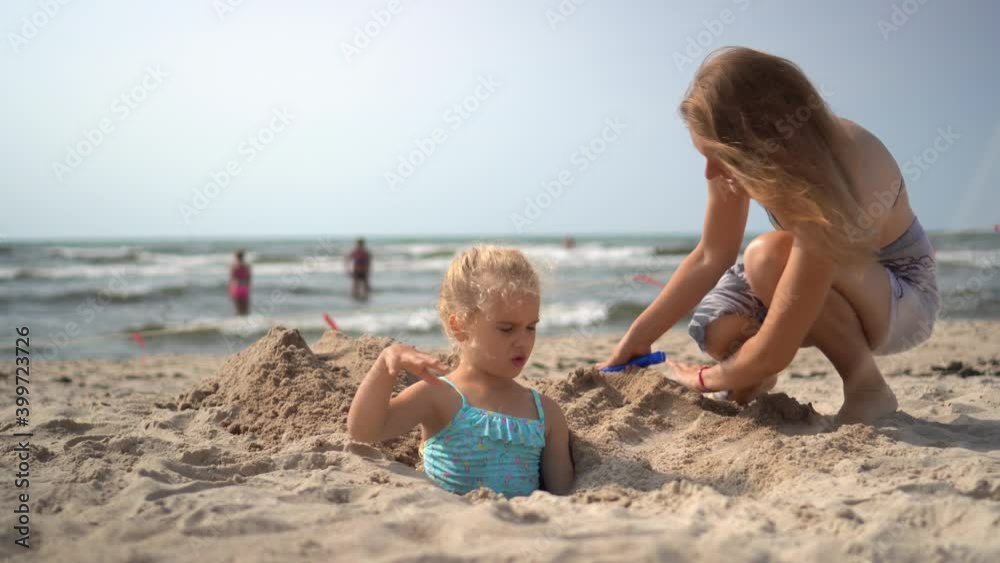 Happy mom bury her little daughter under beach sand. People enjoy summer Happy mom bury her little daughter under beach sand. People enjoy summer