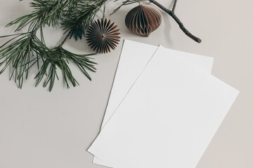 Christmas still life. Blank paper sheets mockups and green pine tree branch on beige table background. Trendy brown Christmas paper ornaments. Winter design, composition. Flat lay, top view.