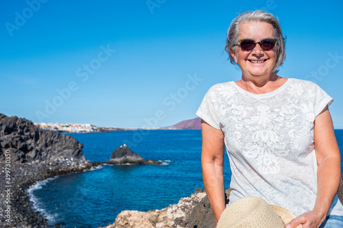 Attractive senior woman white haired standing on the cliffs with sunglasses enjoying vacation or excursion and freedom in seascape. Active retired elderly people
