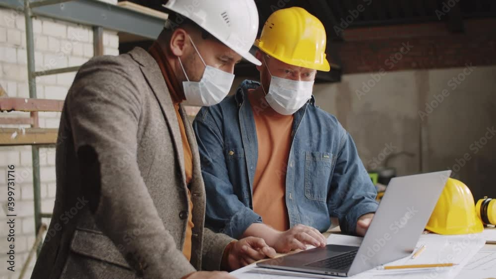 Two male architects in protective face masks and hardhats discussing something on laptop while working together at construction site during coronavirus pandemic