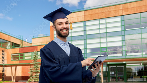 education, graduation and people concept - happy smiling male graduate student in mortar board and bachelor gown with tablet pc computer over school background
