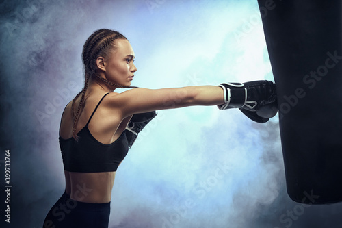 sporty brunette woman in boxing gloves and sportswear kicking bag on dark background with smoke.