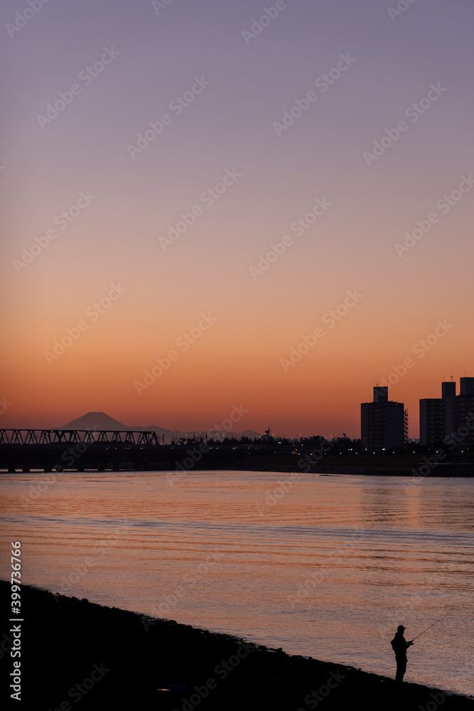 Sunset at Kyuedo River with silhouette of Mount Fuji and Maihama Great Bridge in the background and a person fishing by the river in the foreground.
