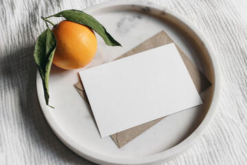 Summer wedding stationery mock-up scene. Blank greeting card, invitation. Craft envelope and orange fruit on elegant marble tray. White muslin tablecloth background. Top view.