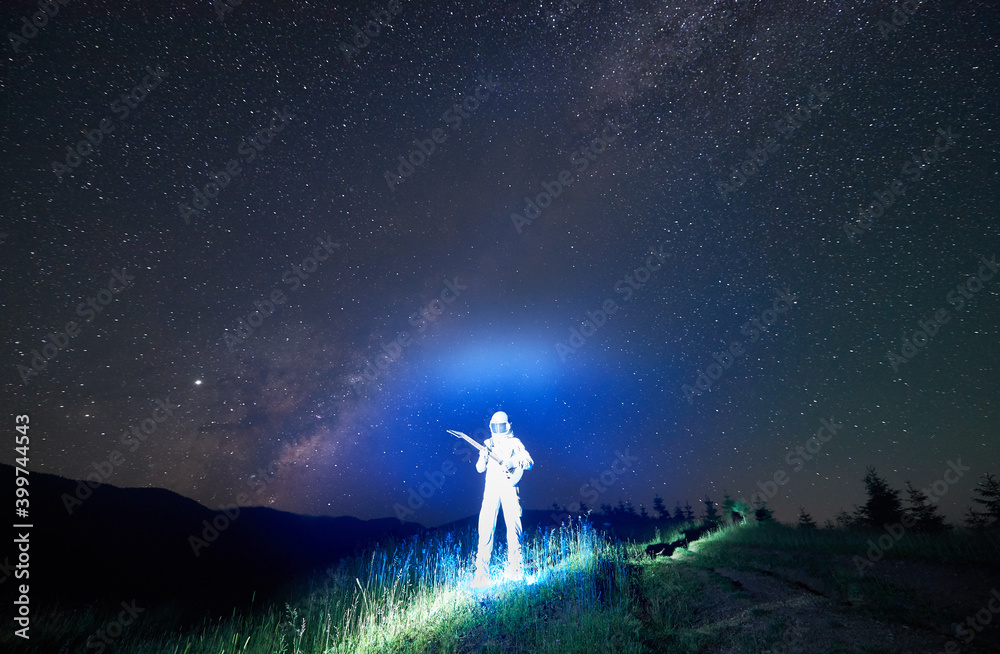Fototapeta premium Illuminated cosmonaut in white space suit and helmet standing in the middle of mountain meadow holding guitar, incredible night sky full of stars on background. Space travel concept